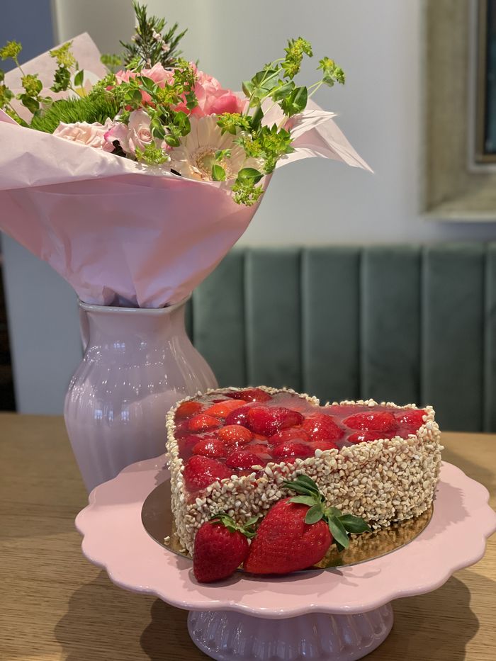Erdbeertorte in Herzform auf einem rosa Tortenständer, daneben ein Blumenstrauß in einer Vase