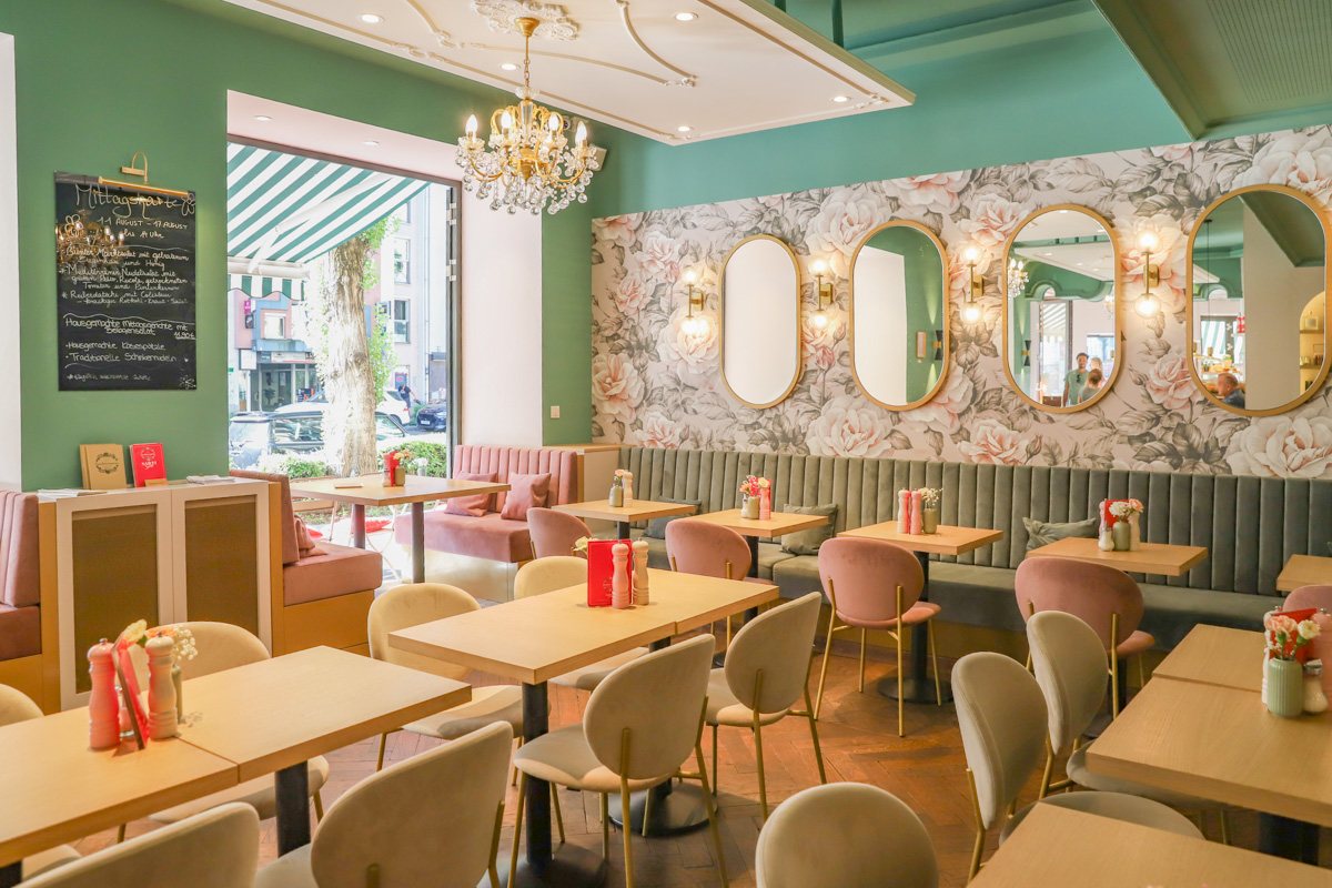 Gastraum des Café Kustermann: helle Tische, rosa Stühle, Kronleuchter und goldgerahmte Spiegel vor Blumentapete