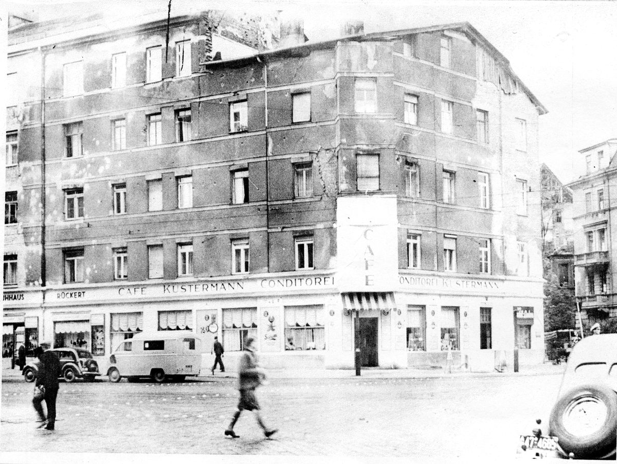 Das Eckhaus mit von Splittern zerschlagener Fassade, davor ein Lieferwagen und Passanten