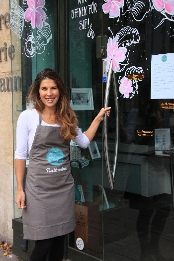 Katharina Stadler lächelnd an der Eingangstür des Cafés, auf dem Glas gemalte Blumen