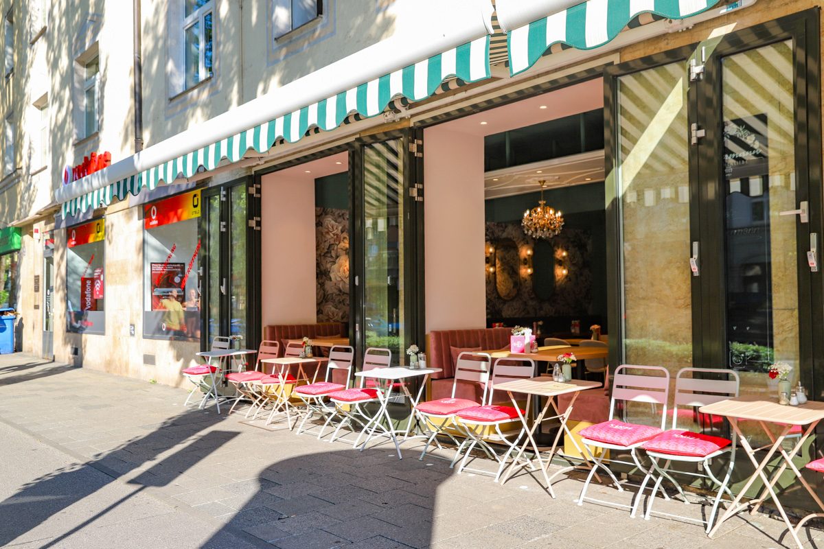 Die Fassade heute mit grün-weiß gestreiften Markisen und Tischen auf dem Gehweg